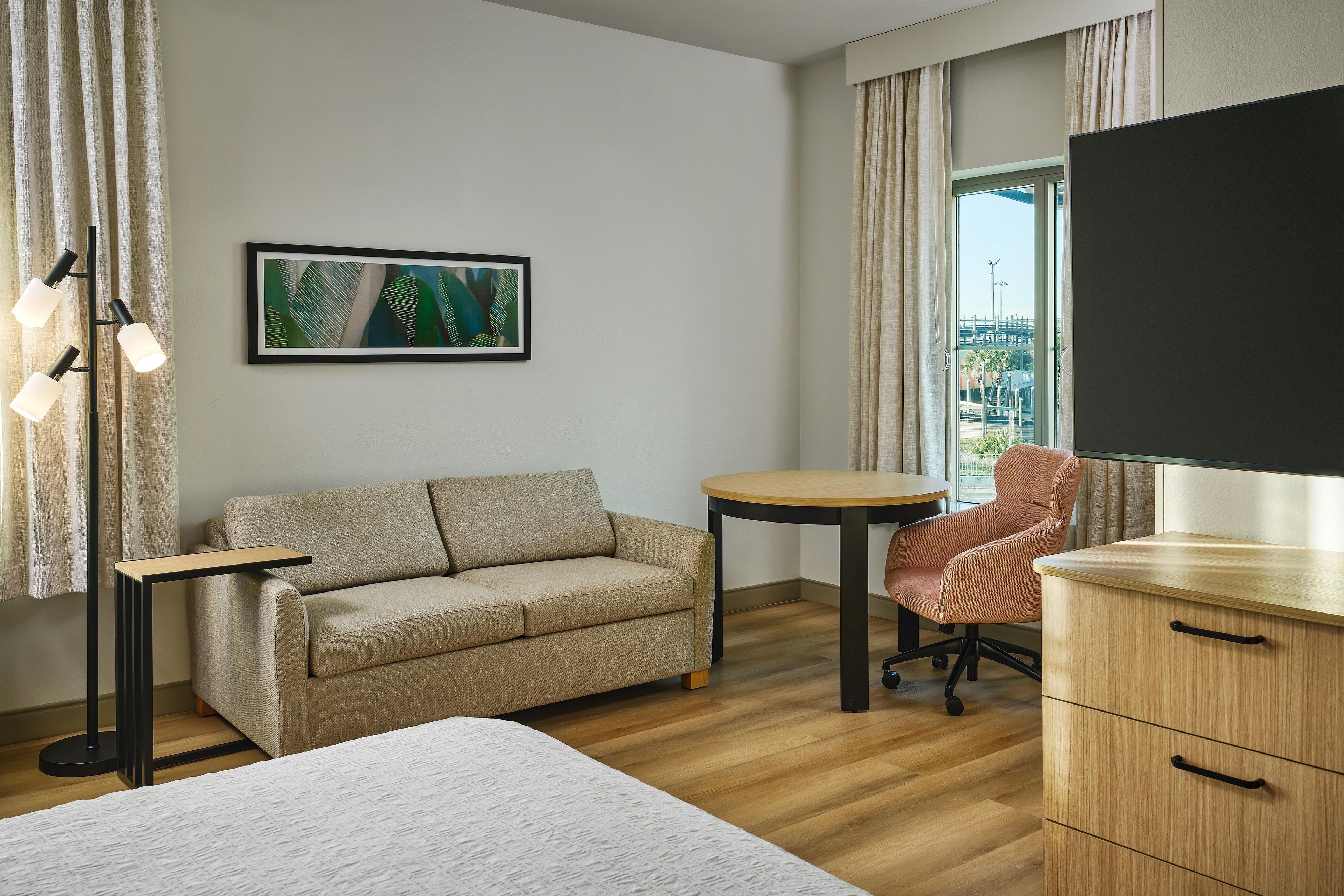 A modern hotel room with a sofa, a small round table, a desk with a chair, and a large window with a view.