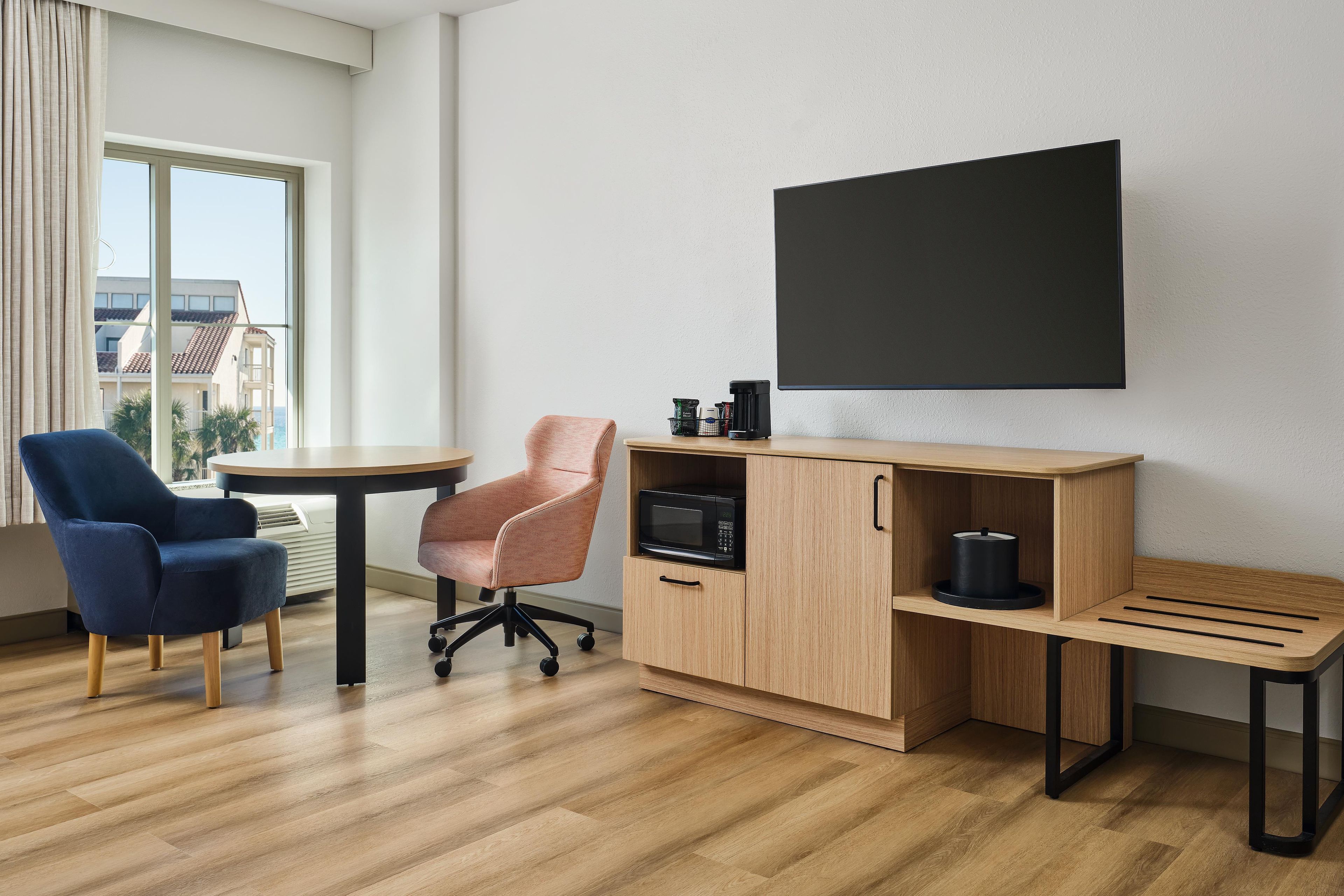 Una habitación moderna con escritorio, dos sillas, cafetera, microondas y un ventanal con cortinas.