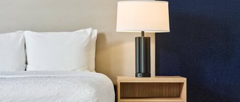 Cama bem arrumada com lençóis brancos, mesa de cabeceira com abajur e parede escura ao fundo.