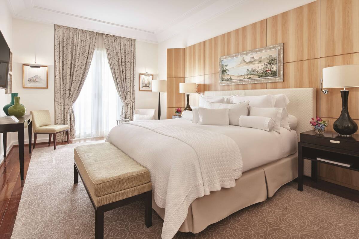 Une chambre d’hôtel avec un grand lit, une table de chevet avec une lampe, une chaise, un bureau et une grande fenêtre avec des rideaux.
