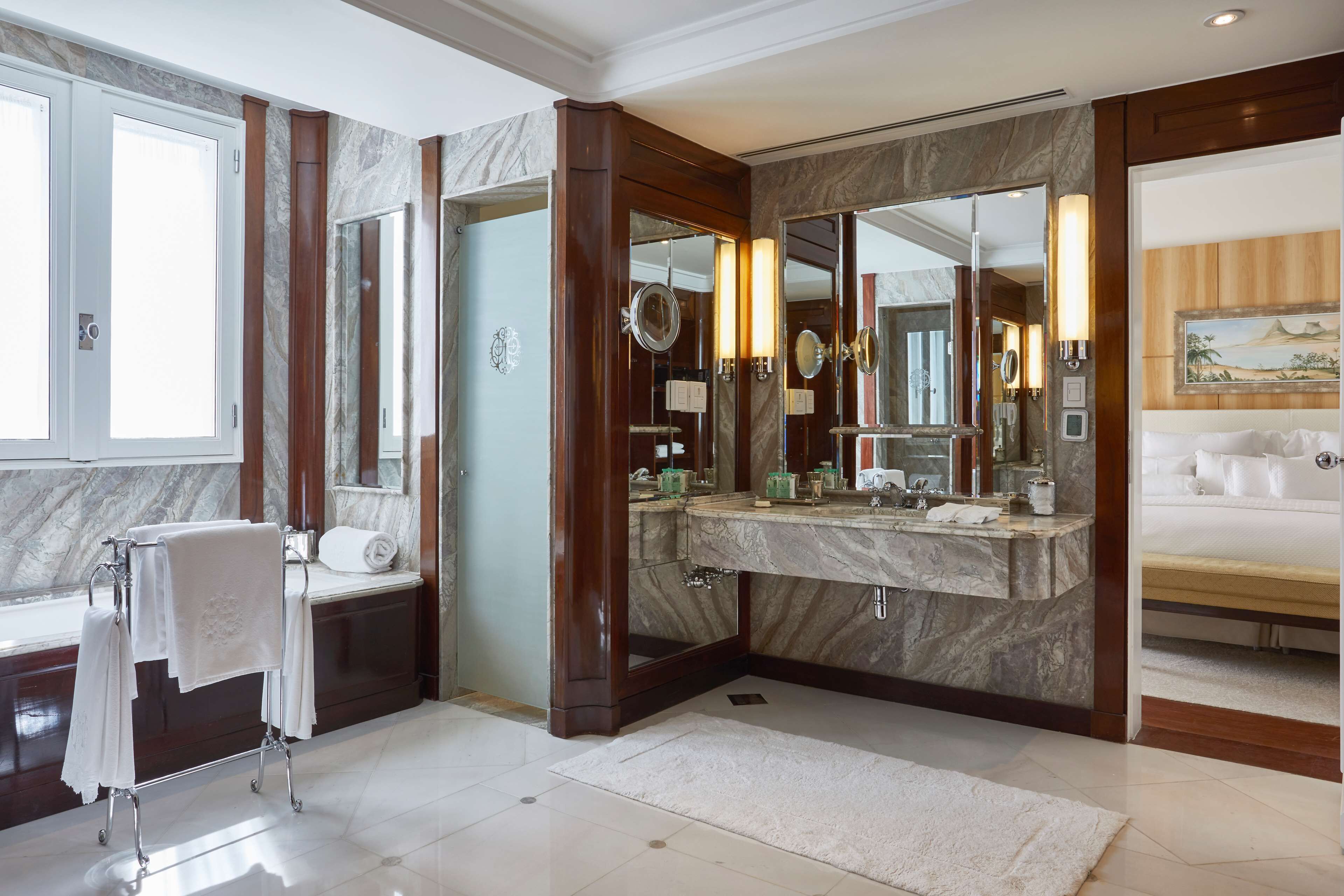 A bathroom with a marble sink, multiple mirrors, a towel rack, and a bed visible through an adjacent room.