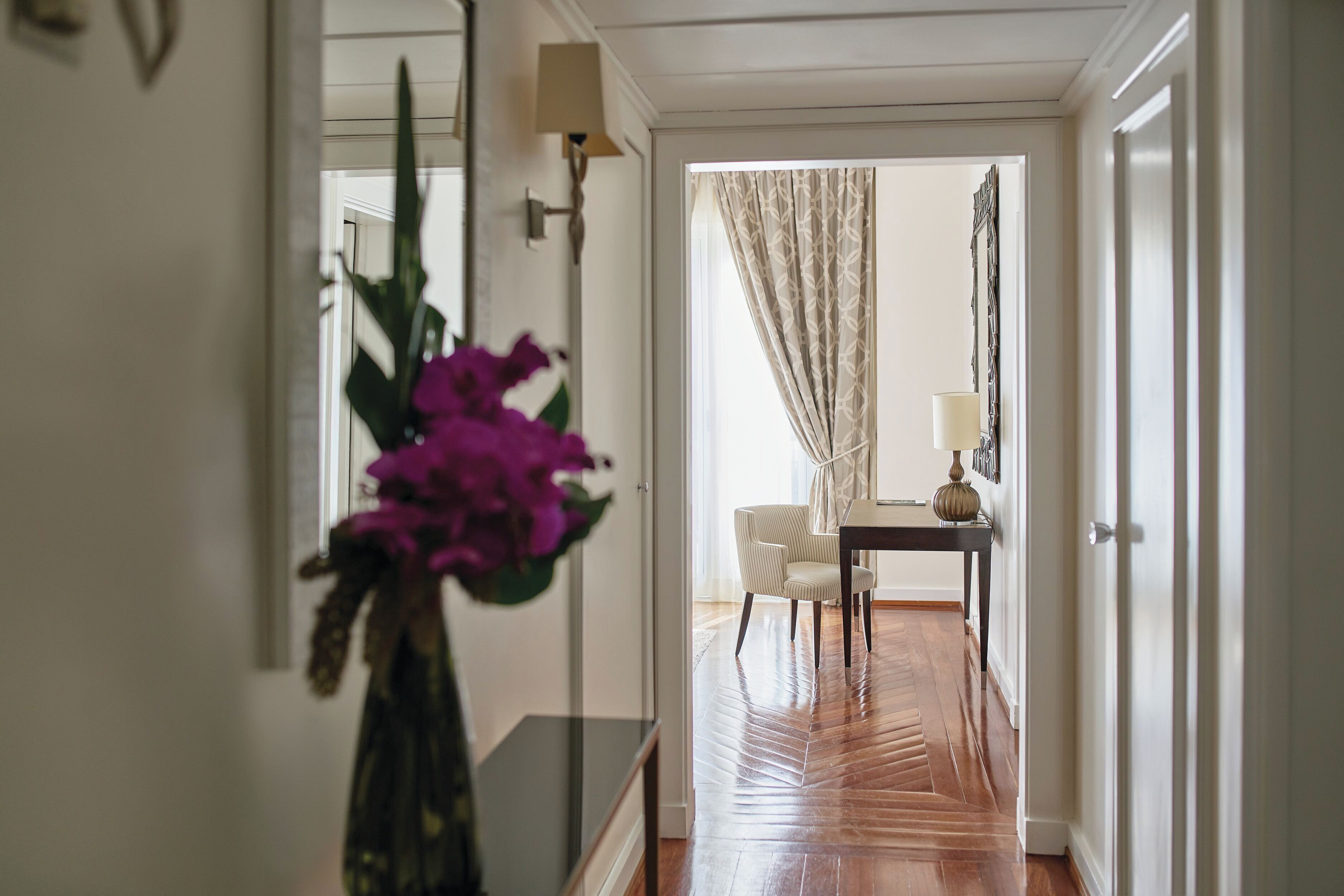 A hallway with a wooden floor, a chair, a table, and a vase with flowers.