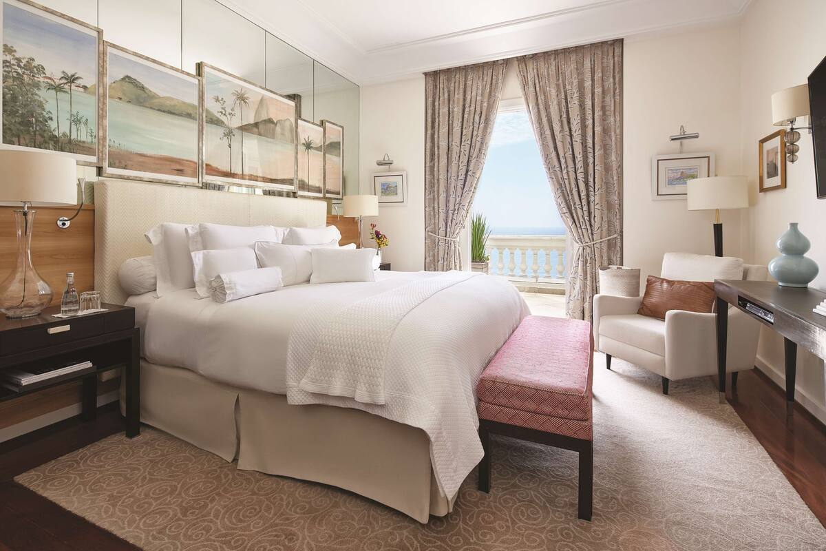 Une chambre d’hôtel avec un grand lit, un bureau, une chaise, une petite table, une lampe, un miroir et une vue sur la mer.