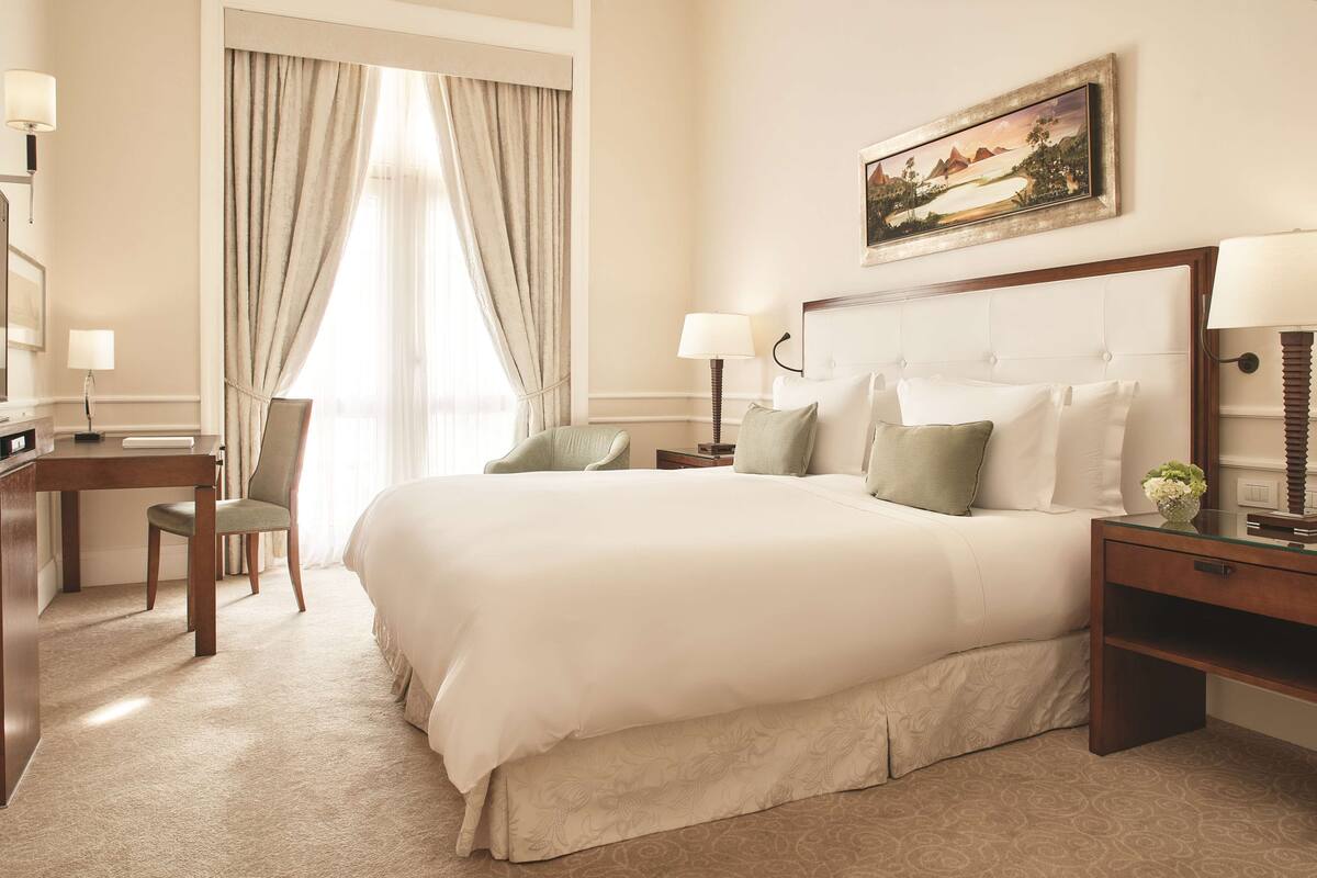 Une chambre d’hôtel avec un grand lit, deux tables de chevet, un bureau avec une chaise, une fenêtre avec des rideaux et un tableau au mur.