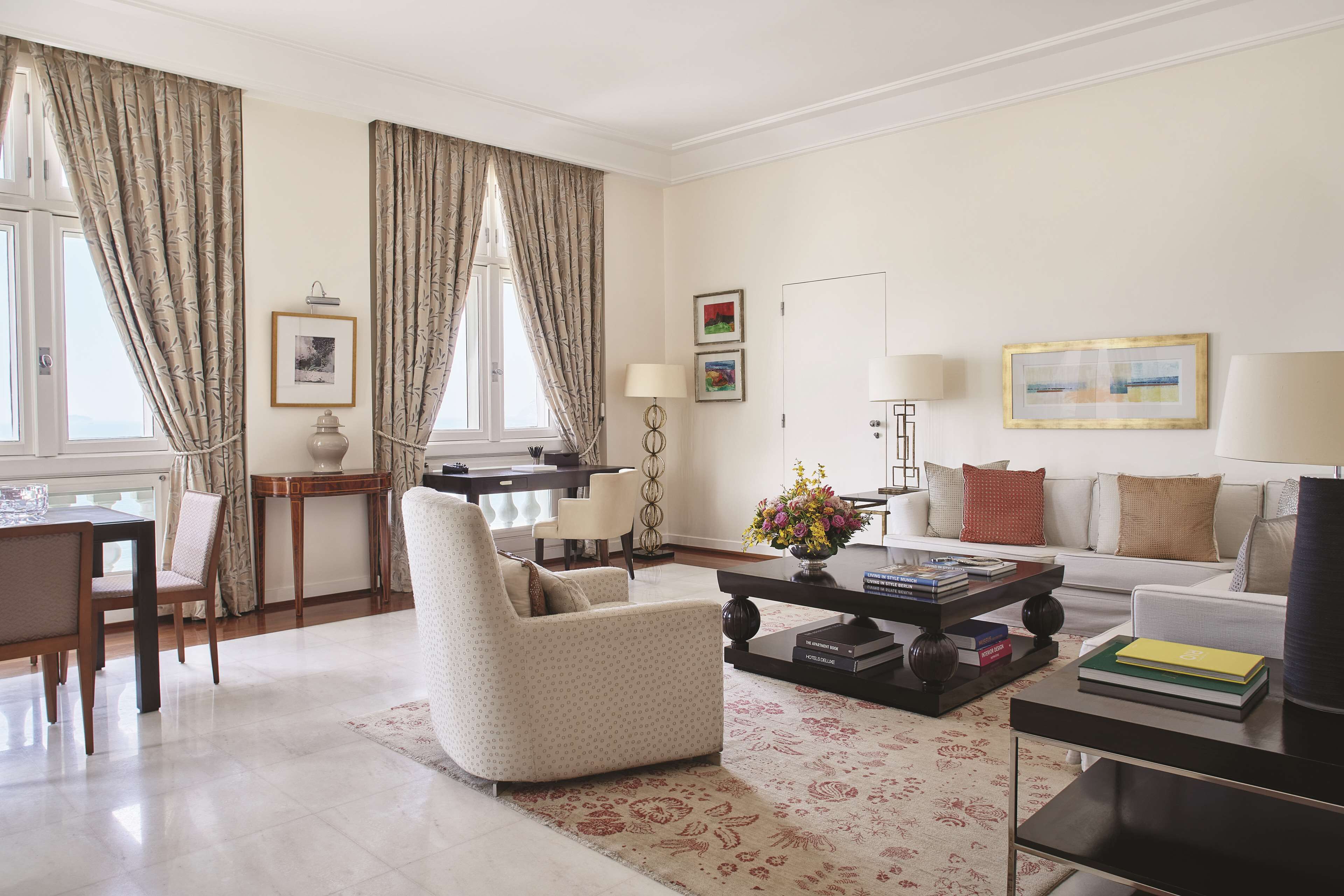 A well-lit living room with a sofa, armchair, dining table, and coffee table. There are curtains, framed pictures on the walls, and a vase with flowers on the coffee table.