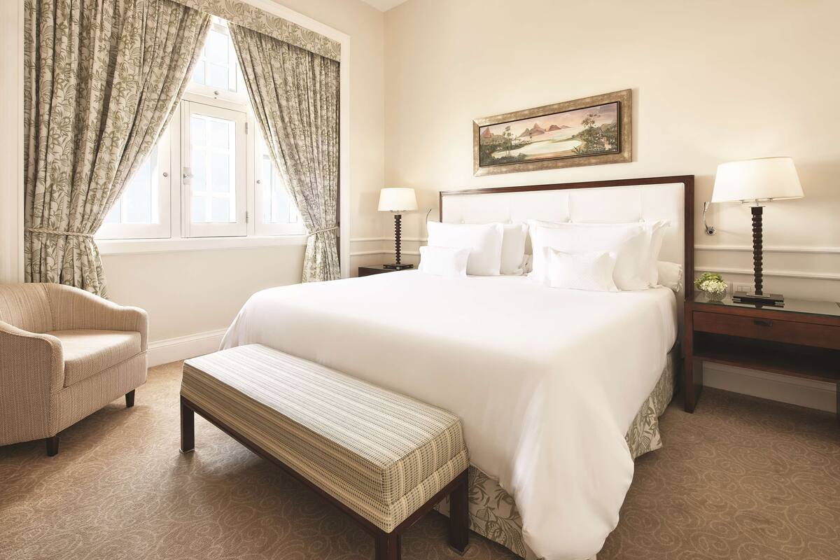 Une chambre d’hôtel bien aménagée, avec un grand lit, une banquette, une chaise, deux tables de chevet avec des lampes et un tableau au mur.