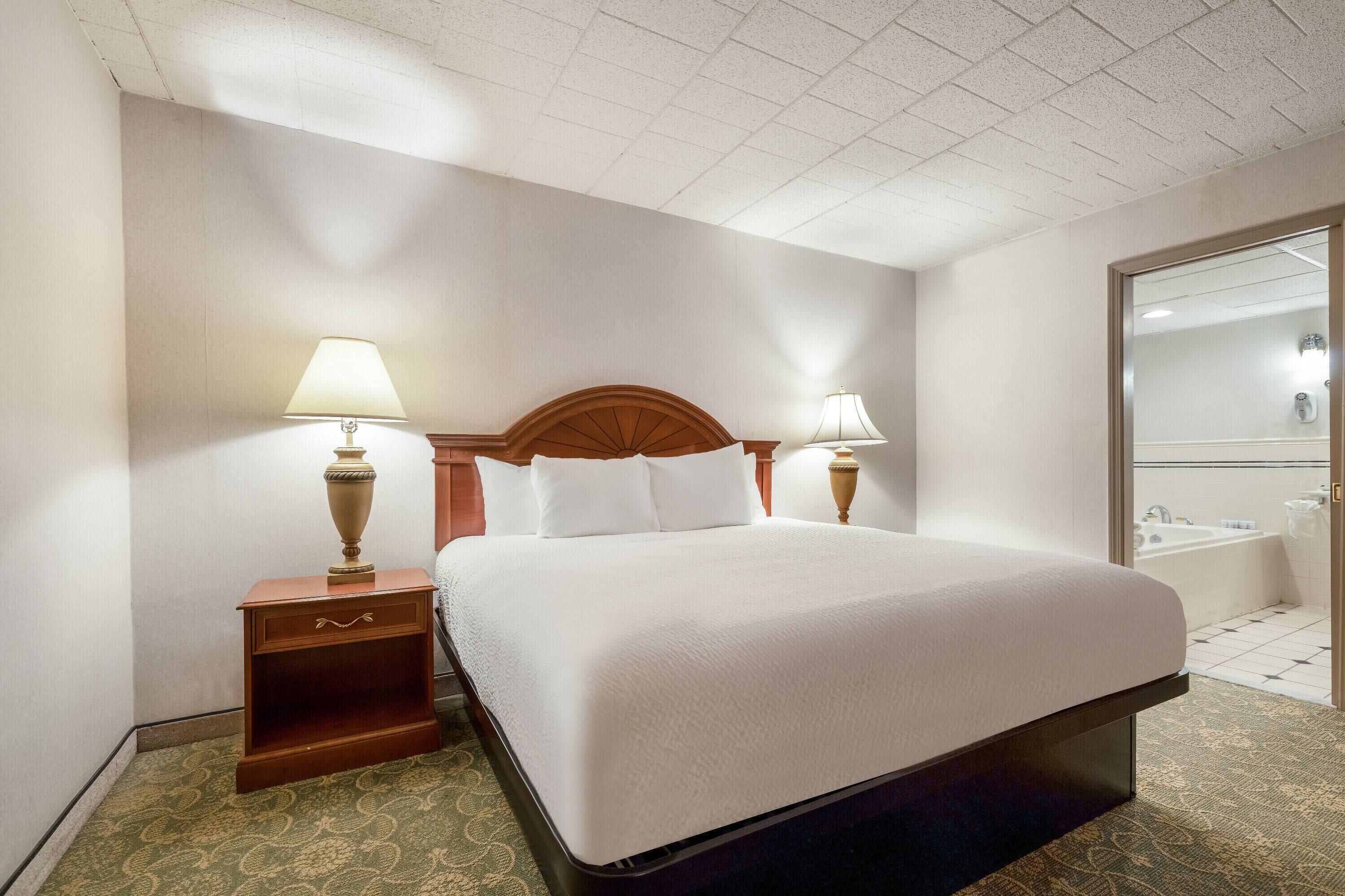 Ein Hotelzimmer mit einem großen Bett, zwei Nachttischlampen, einem hölzernen Kopfteil, einem kleinen Nachtisch und einem durchgegangenen Tür erkennbaren Badezimmer.