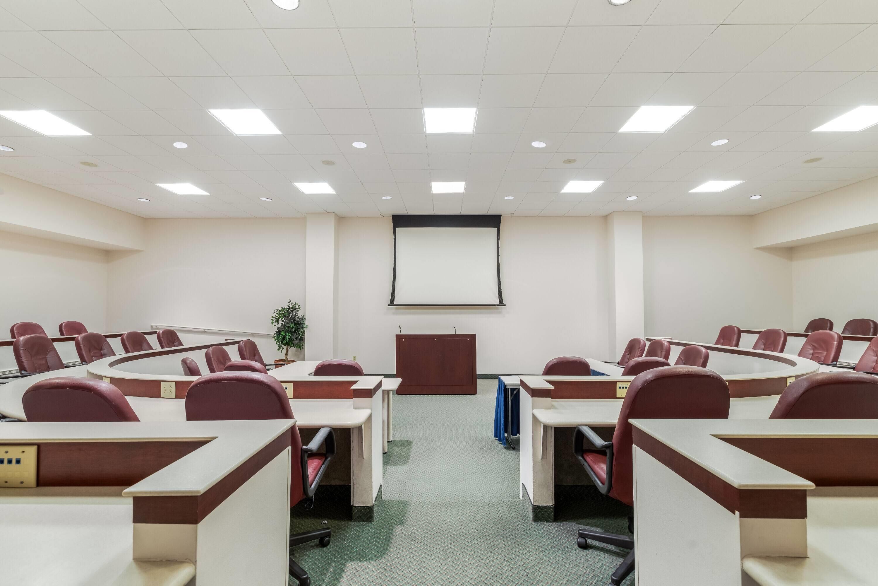 A conference room with a large screen, multiple tables, and chairs arranged in a semi-circular layout.