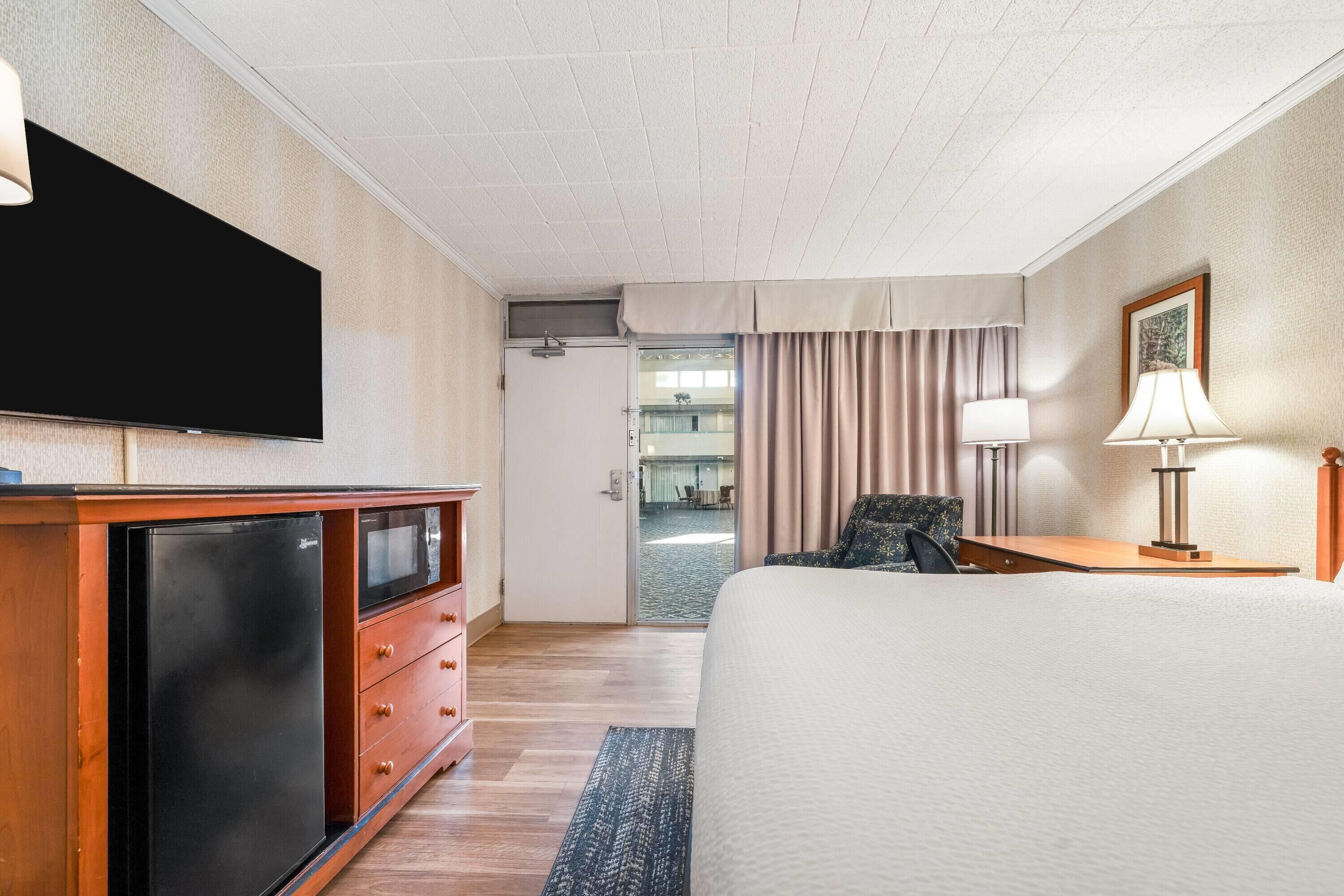 Habitación de hotel con cama, televisor de pantalla plana, refrigerador, escritorio con lámpara y vista al pasillo y otra habitación.