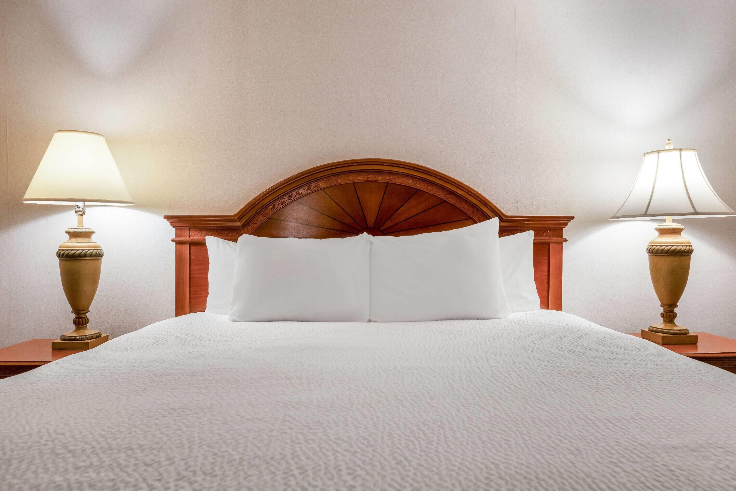 A neatly made bed with white linens, flanked by two bedside tables with lamps, in a room with a wooden headboard and plain walls.