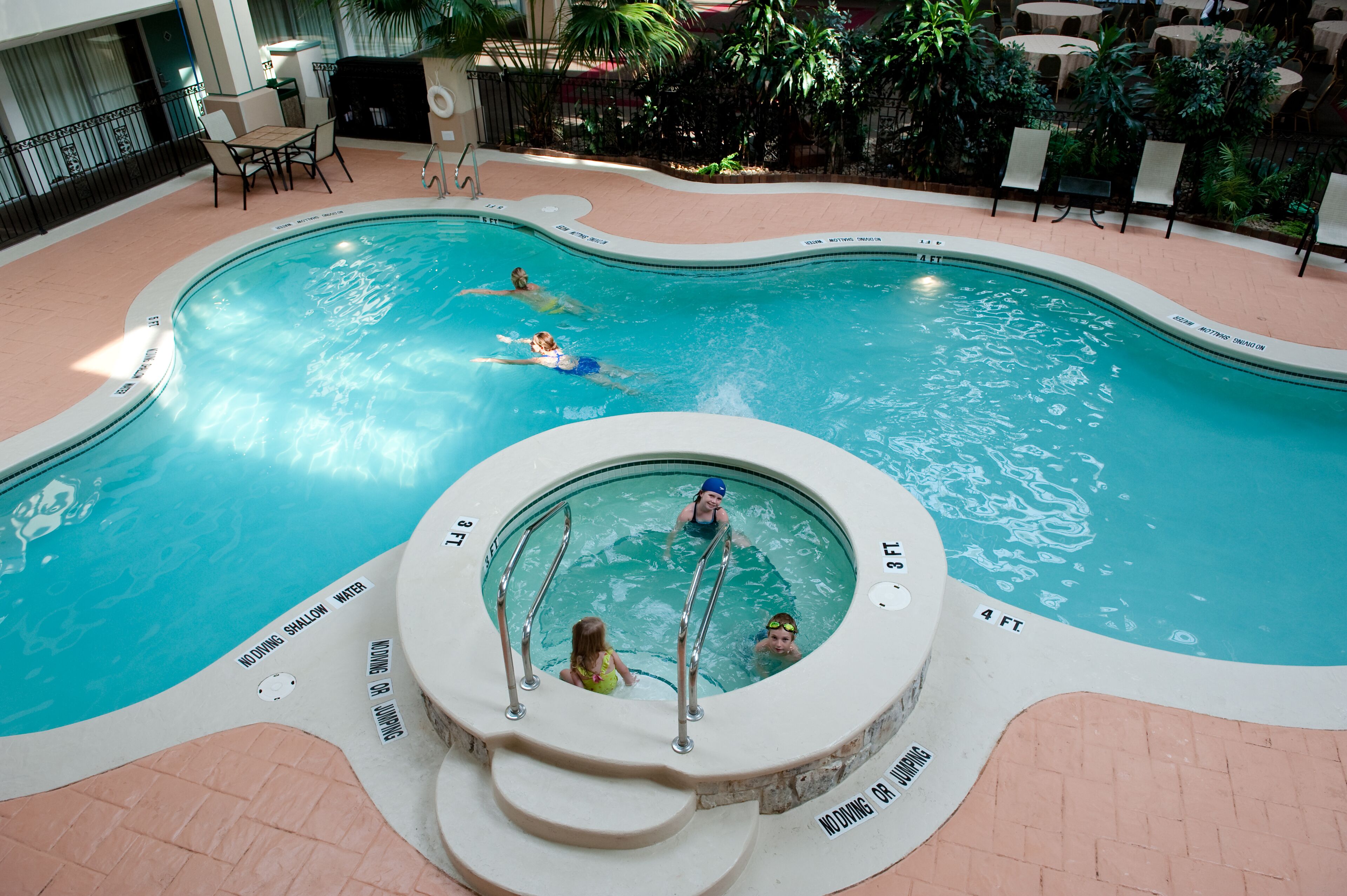 An indoor swimming pool with a hot tub, lounge chairs, and a patio area with tables and chairs.