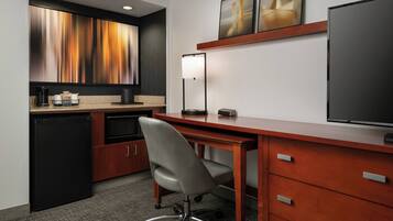 A compact hotel room with a kitchenette, a desk with a chair, a flat-screen TV, and artwork on the wall.