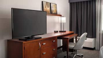 Hotelkamer met een houten bureau, een flatscreen televisie, een lamp, een stoel en een groot raam met gordijnen.