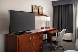 A hotel room with a wooden desk, a flat-screen TV, a lamp, a chair, and a large window with curtains. - Courtyard Fremont Silicon Valley (Fremont)