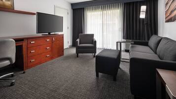 A hotel room with a flat-screen TV, a sofa, a chair, a desk with drawers, and a large window with curtains.