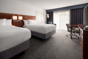 A hotel room with two beds, a desk, a chair, and a large window with curtains.