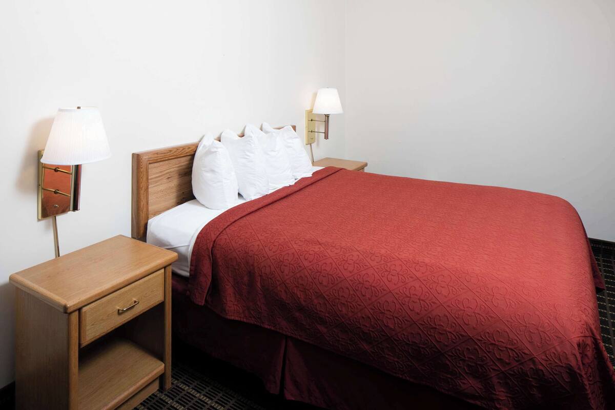 Ein Hotelzimmer mit einem hölzernen Kopfteil, einer roten Bettdecke, zwei Nachttischen mit Lampen und einer Schublade.