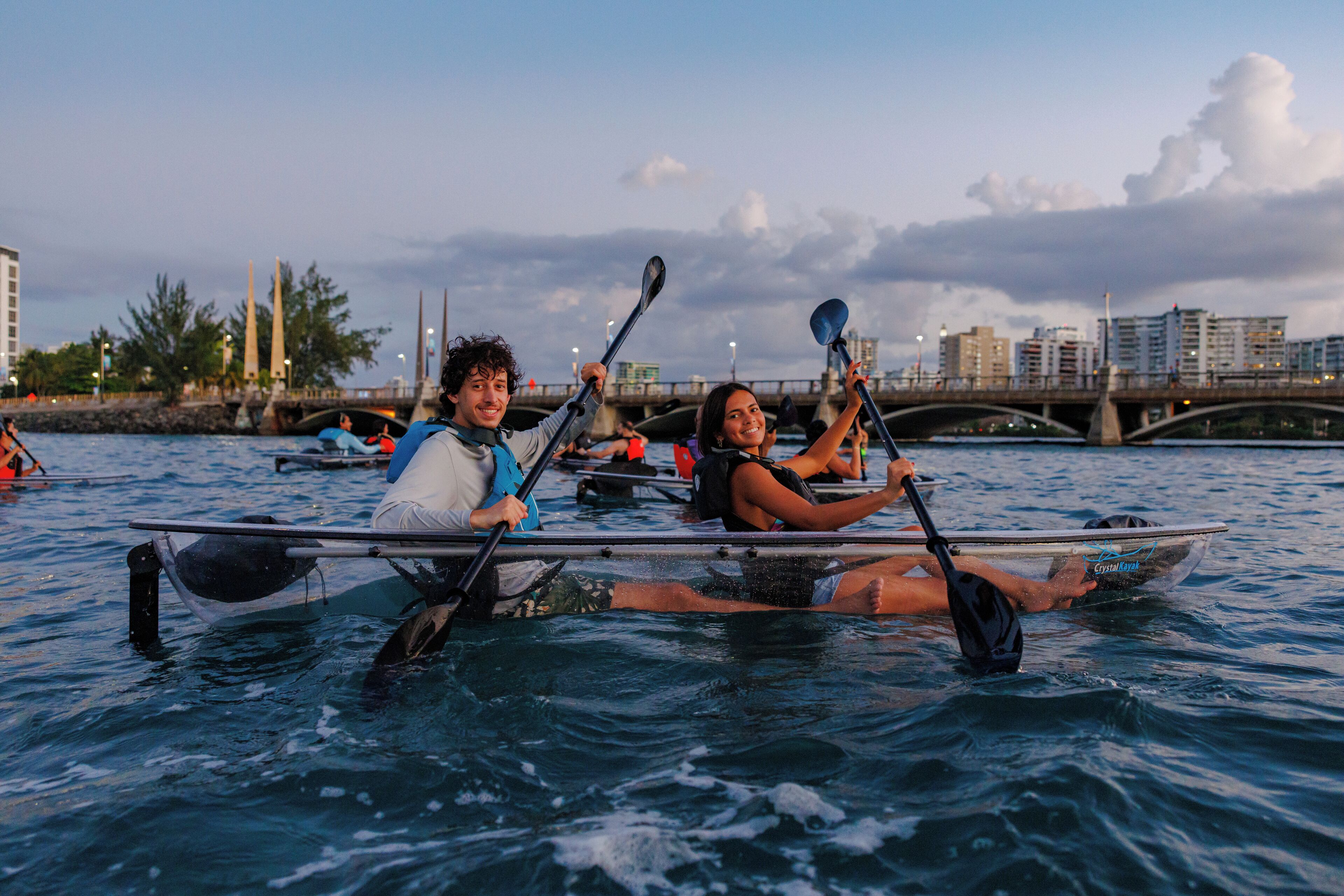 Due persone su un kayak in mezzo all'acqua, con un ponte e degli edifici sullo sfondo.