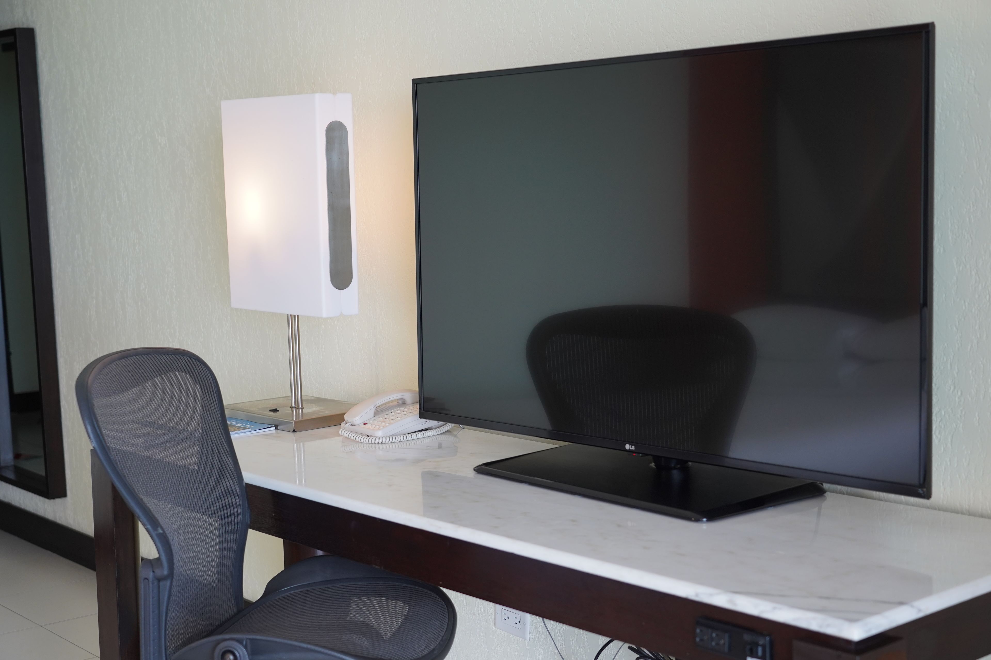 A hotel room with a large flat-screen TV, a desk with a chair, a lamp, and a telephone.