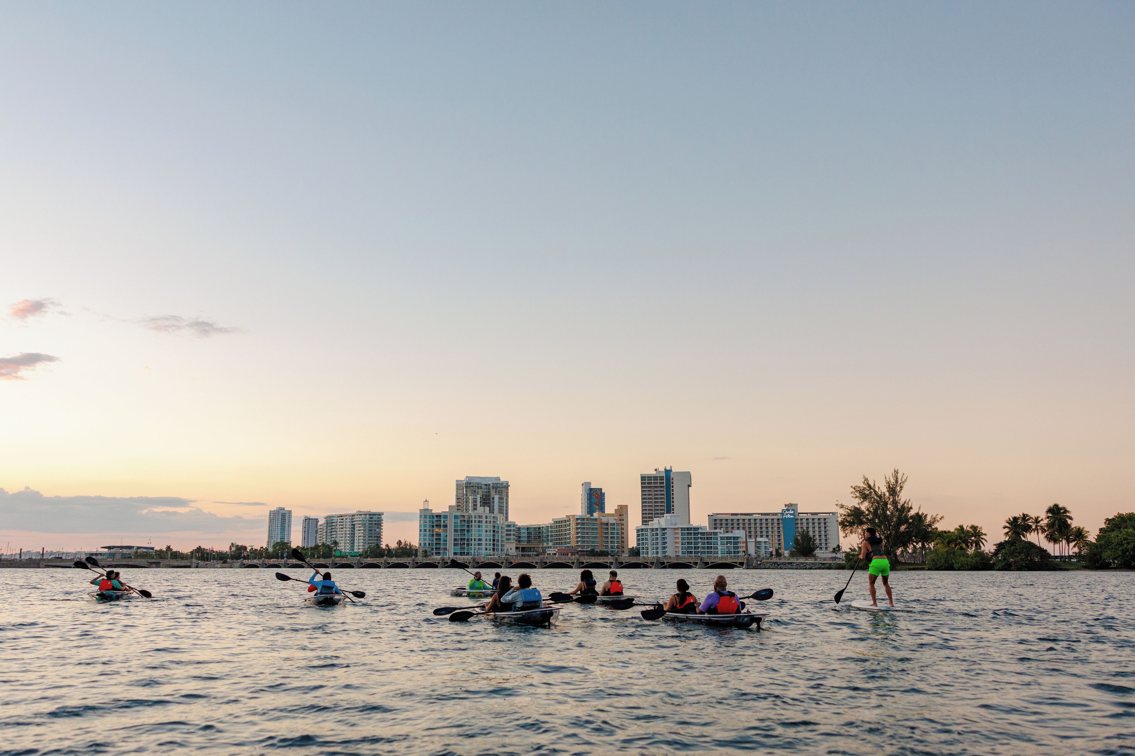 A group of people kayaking on a calm body of water with a city skyline in the background.