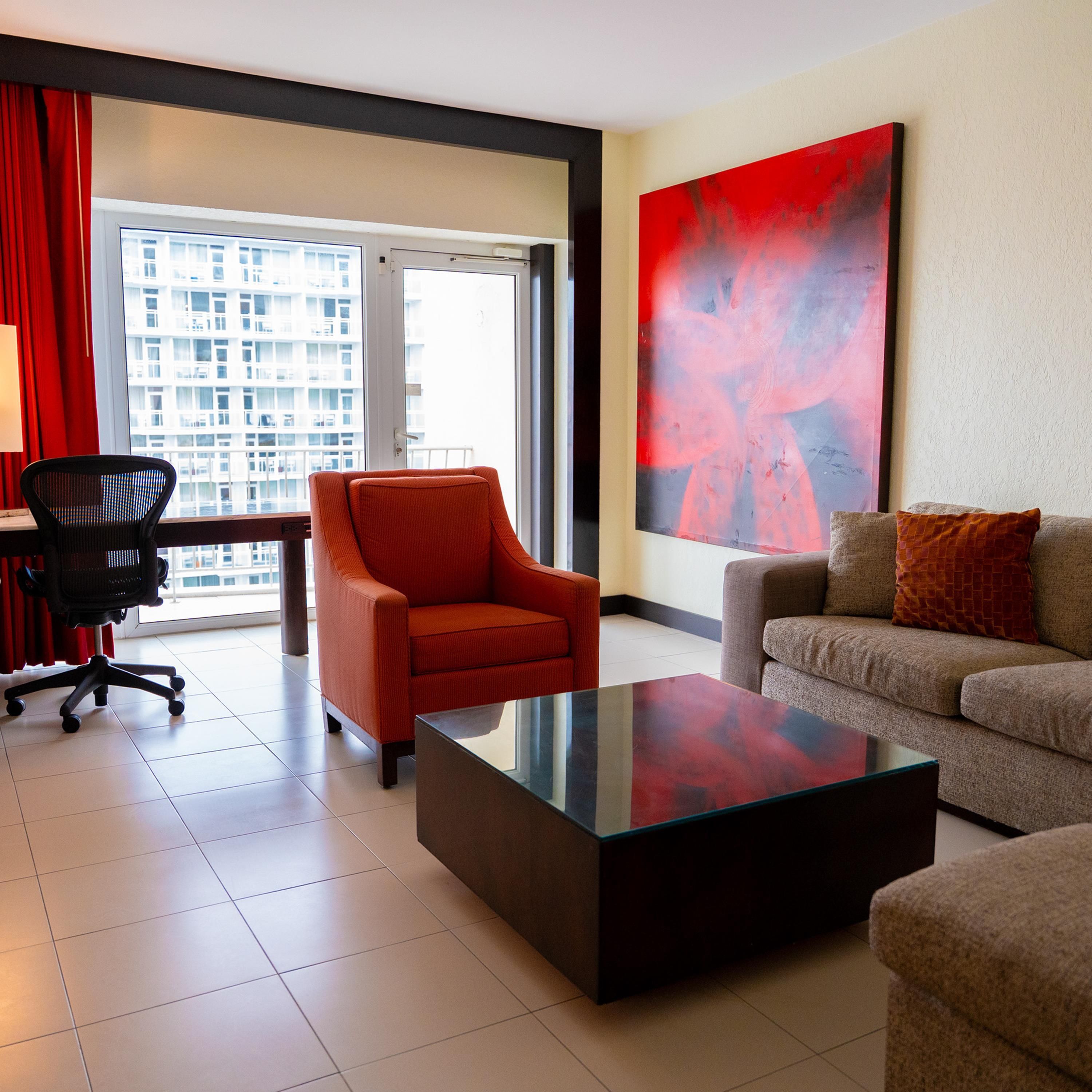Un hotel moderno con una ventana grande, un sillón rojo, un sofá, una mesa de centro de cristal y un cuadro abstracto de gran tamaño en la pared.