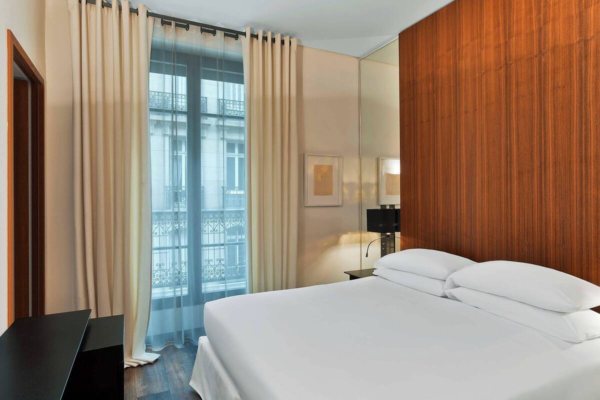 A hotel room with a large bed, a wooden panel wall, a window with curtains, and a nightstand with a lamp.