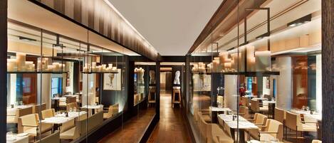A modern restaurant with long tables and chairs, glass partitions, and warm lighting.