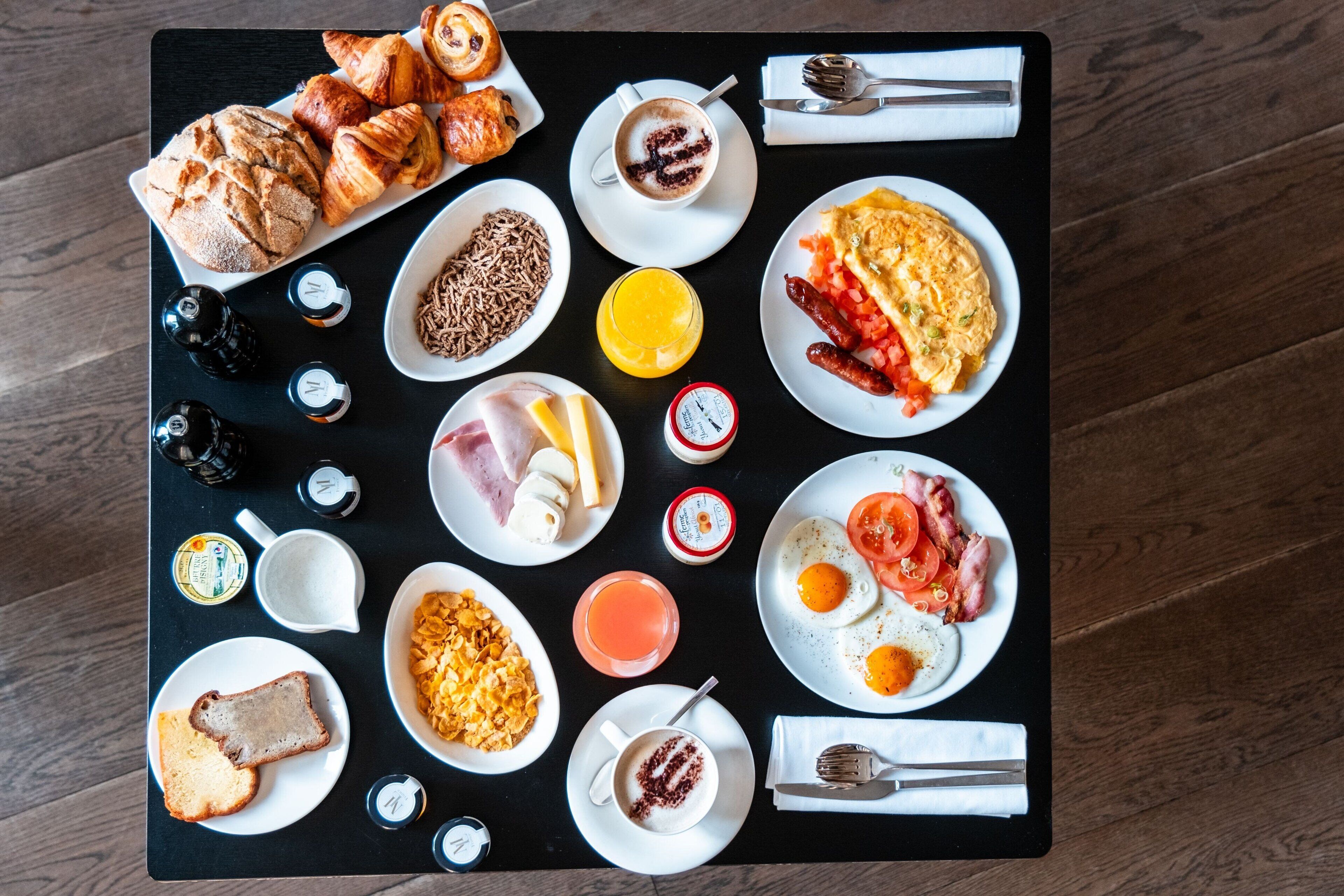 Un desayuno servido en una bandeja negra con cruasanes, huevos, salchichas y diferentes condimentos.