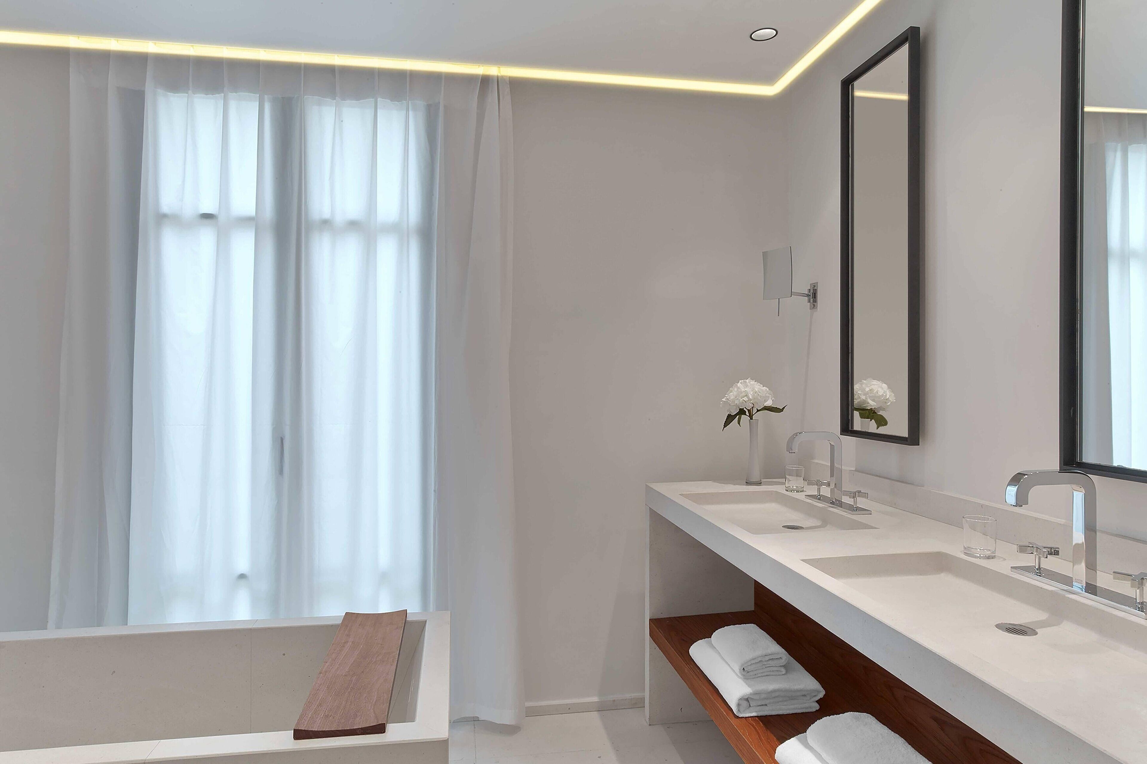 A modern bathroom with two sinks, a large mirror, and a window with sheer curtains.
