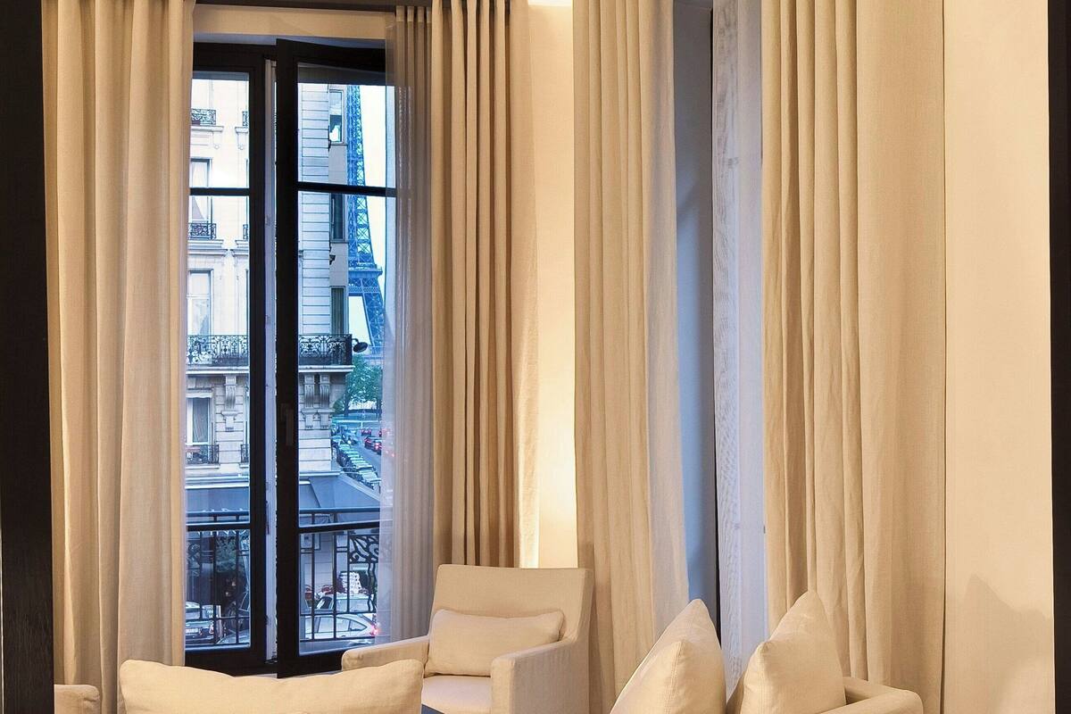 A hotel room with a bed, a sofa, and a chair. There is a large window with curtains, and a view of buildings outside.