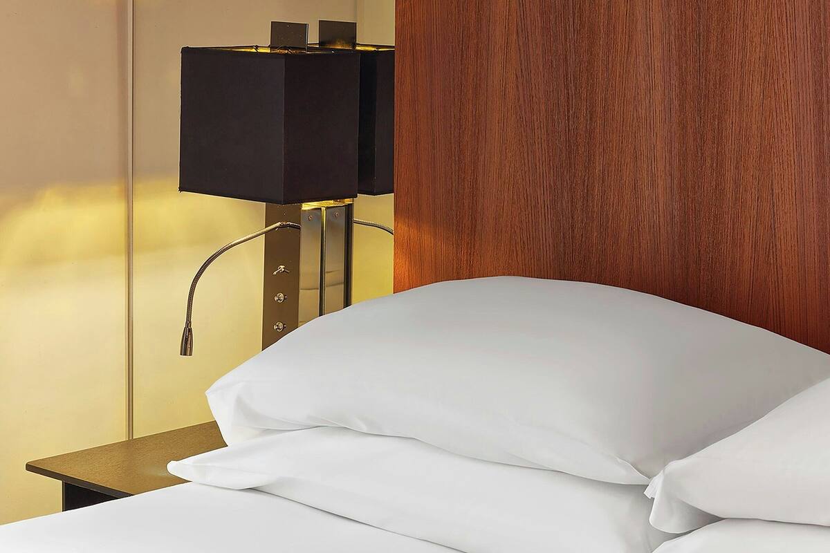 A hotel room with a bed, white pillows, a nightstand, a lamp, and a wooden panel wall.