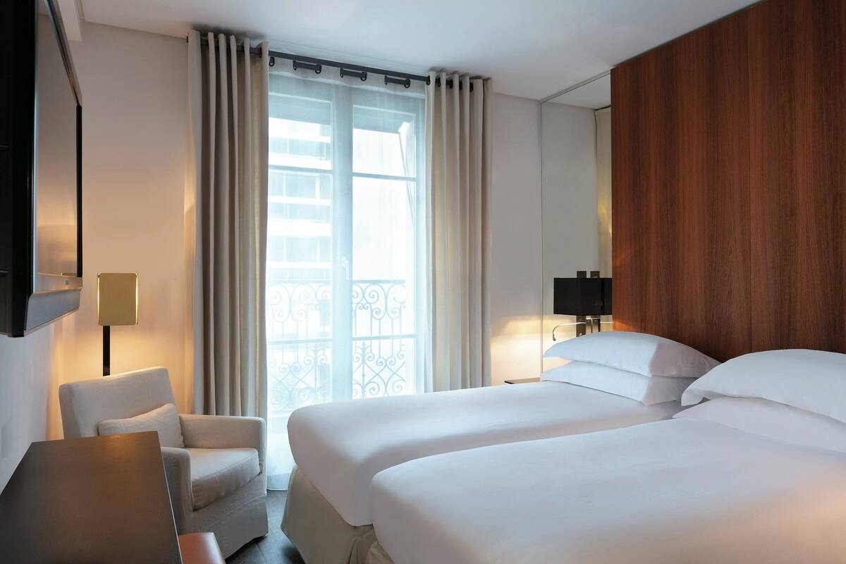 A hotel room with two beds, a chair, a desk, and a large window with curtains.