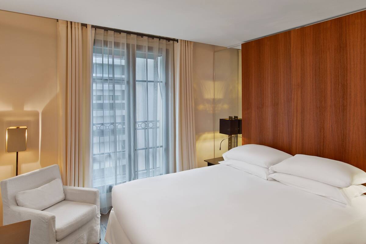 A hotel room with a large bed, a chair, a nightstand, and a window with curtains.