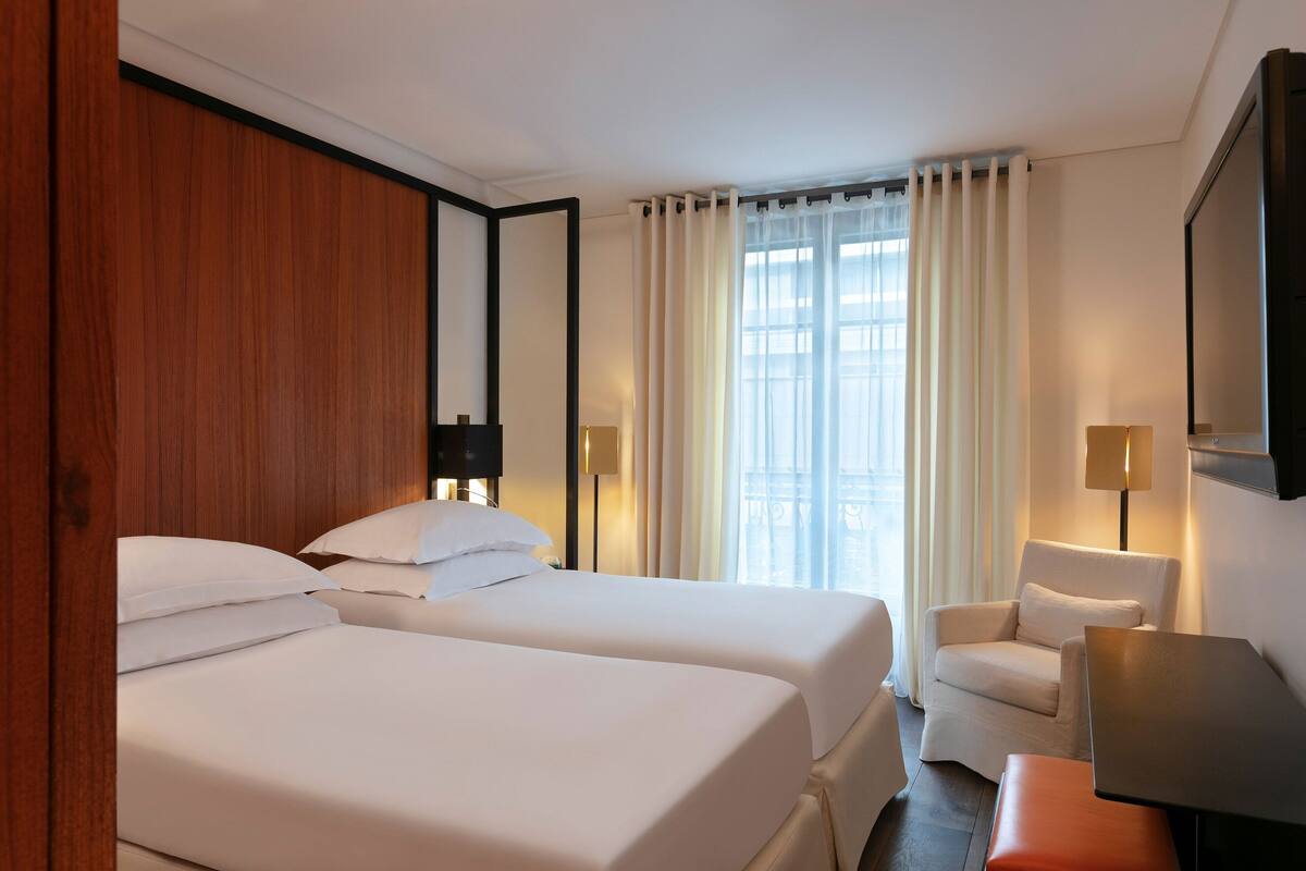 A hotel room with two beds, a chair, a table, and a large window with curtains.