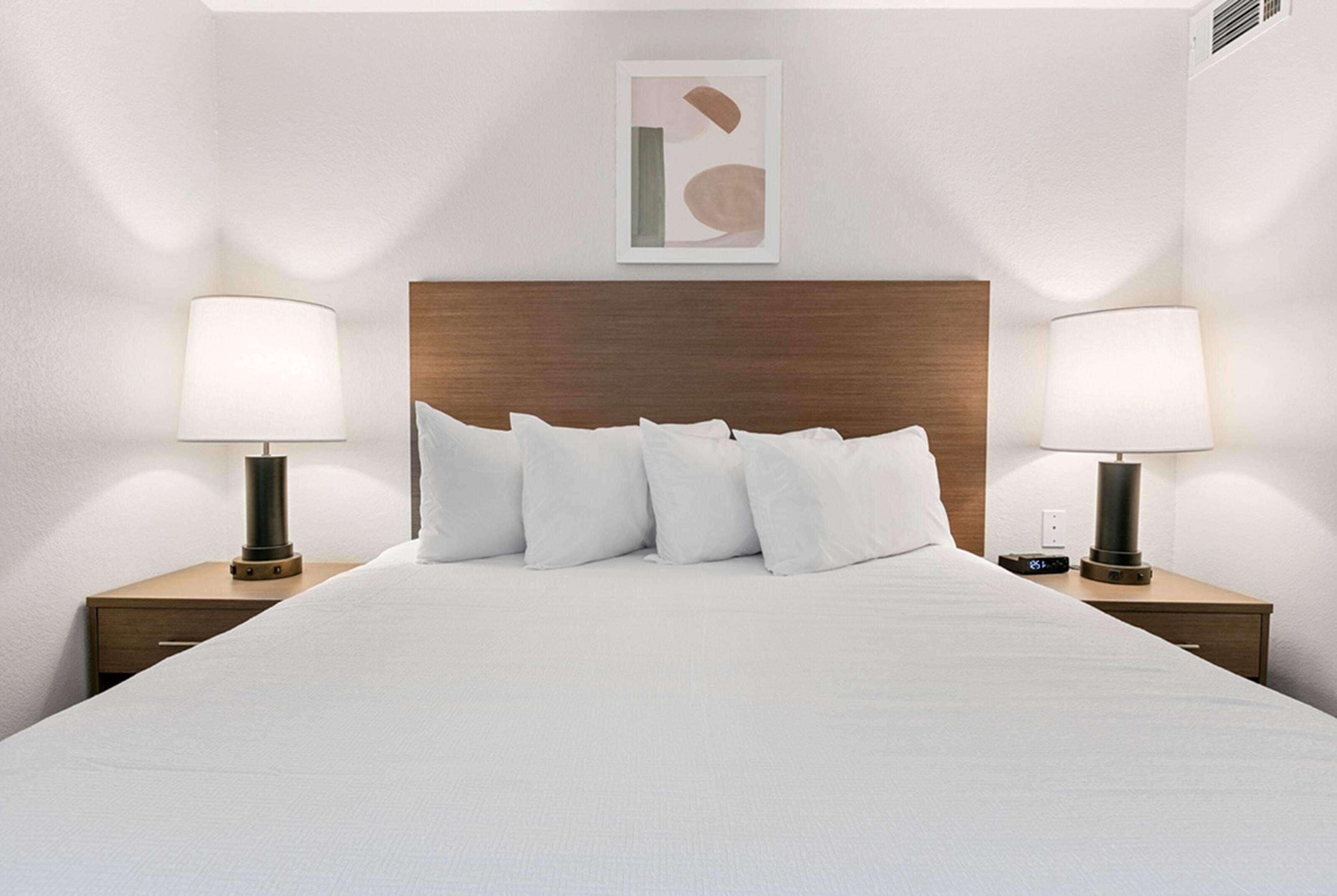 Una cama bien hecha con sábanas blancas y un cabecero de madera. Hay dos mesitas de noche, con lámparas y un pequeño dispositivo electrónico sobre una de ellas.