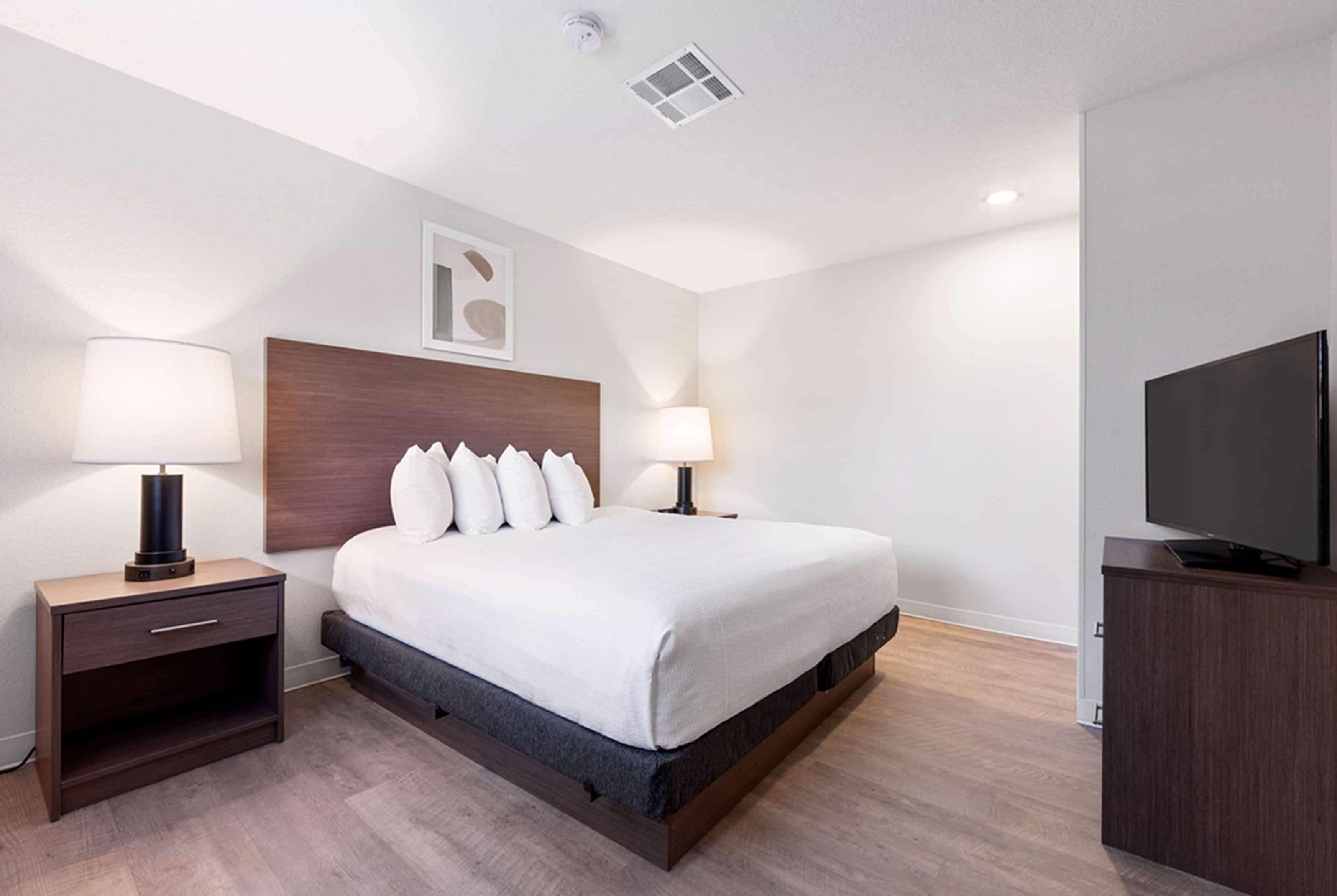 A modern hotel room with a large bed, bedside tables with lamps, a flat-screen TV, and a ceiling vent.