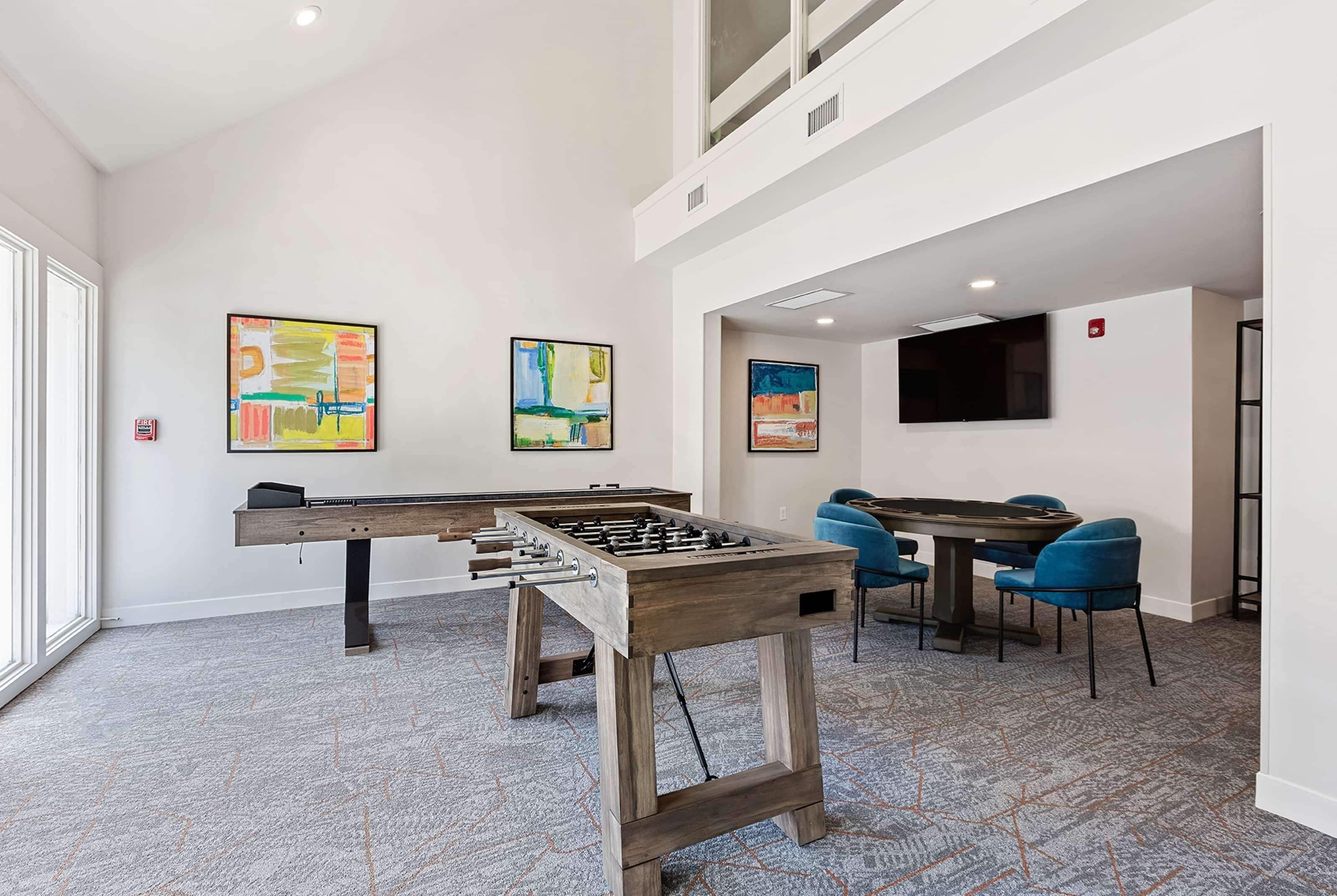 Una habitación con mesa de fútbol, mesa de billar y zona de comedor con sillas azules. Hay pinturas abstractas en las paredes y un televisor de pantalla plana montado sobre la mesa de comedor.