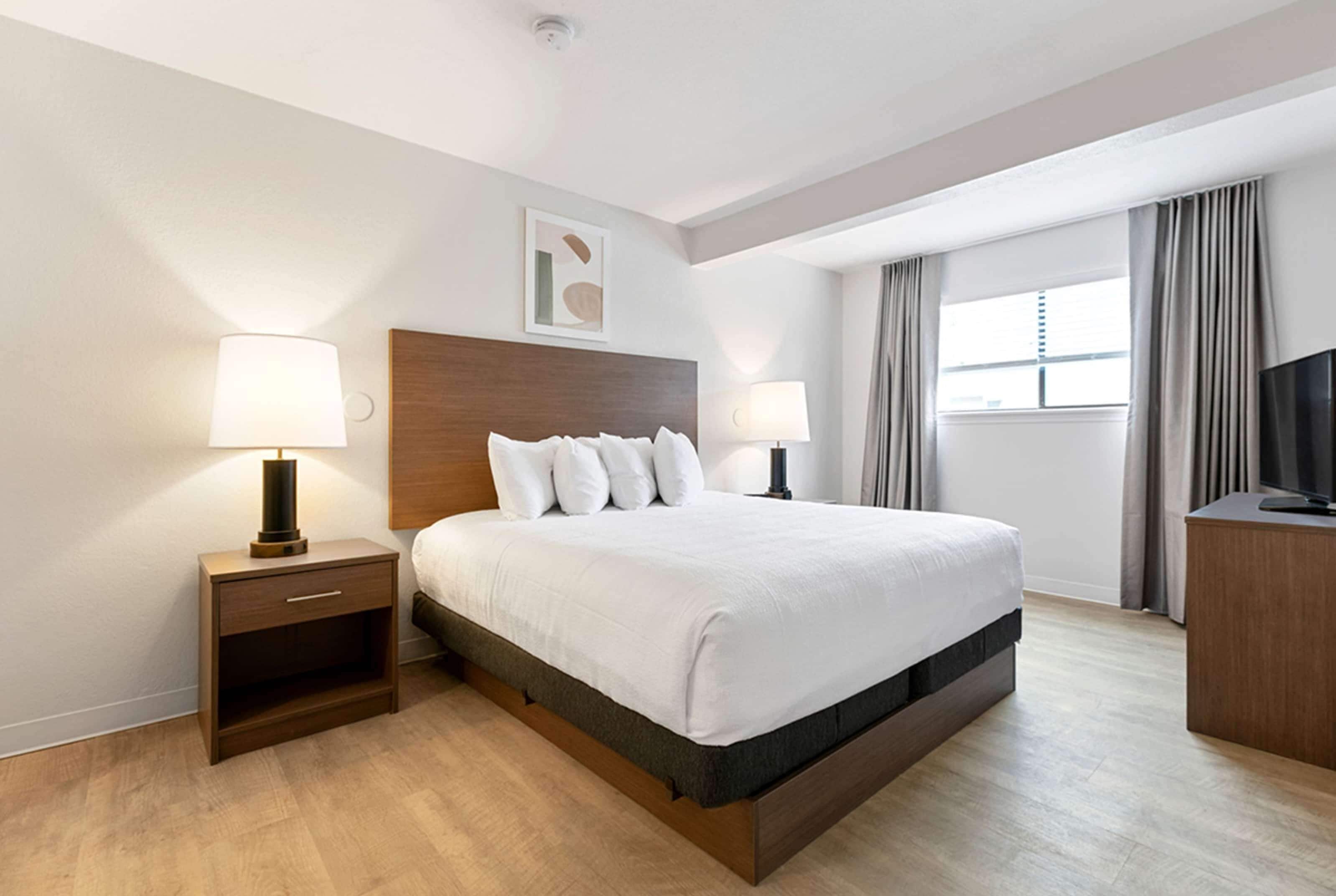 A modern hotel room with a large bed, two bedside tables with lamps, a flat-screen TV, and a window with curtains.