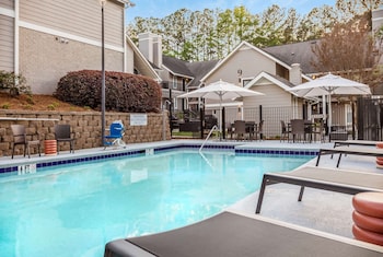 A modern library with a dark wood bookshelf filled with books, a central desk with chairs, and a view of a dining area through large windows. at WaterWalk Extended Stay by Wyndham Atlanta Perimeter