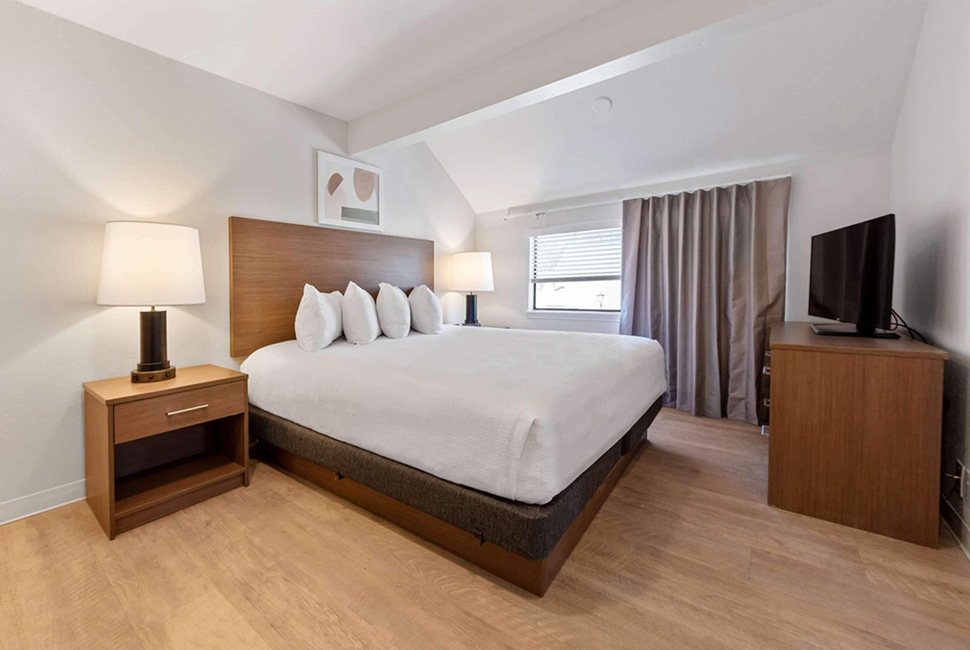Quarto de hotel com uma cama grande, duas mesas de cabeceira com luminárias, uma televisão, uma janela com cortinas e uma luz no teto.