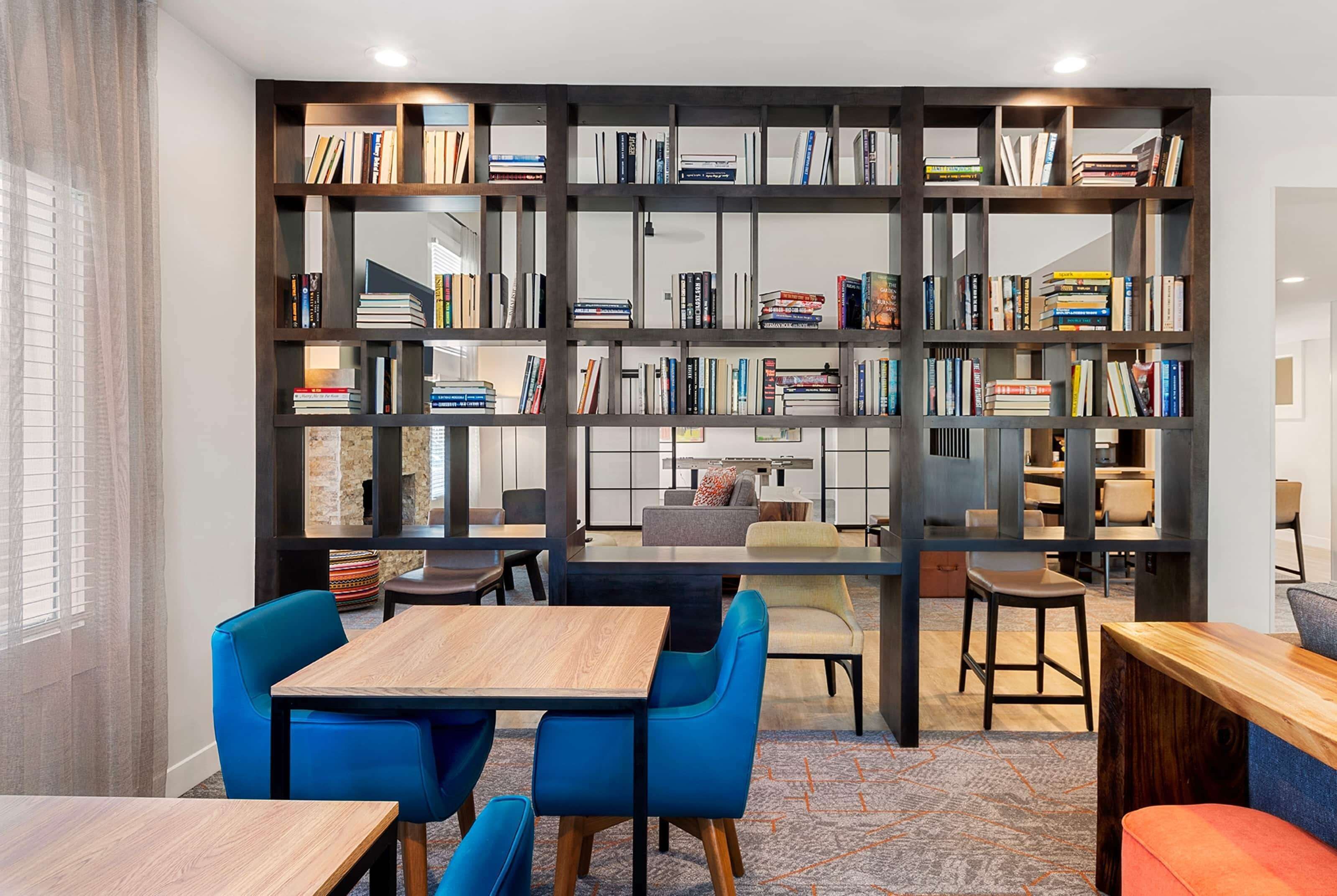 A modern library with bookshelves, a wooden table, blue chairs, and a patterned carpet.