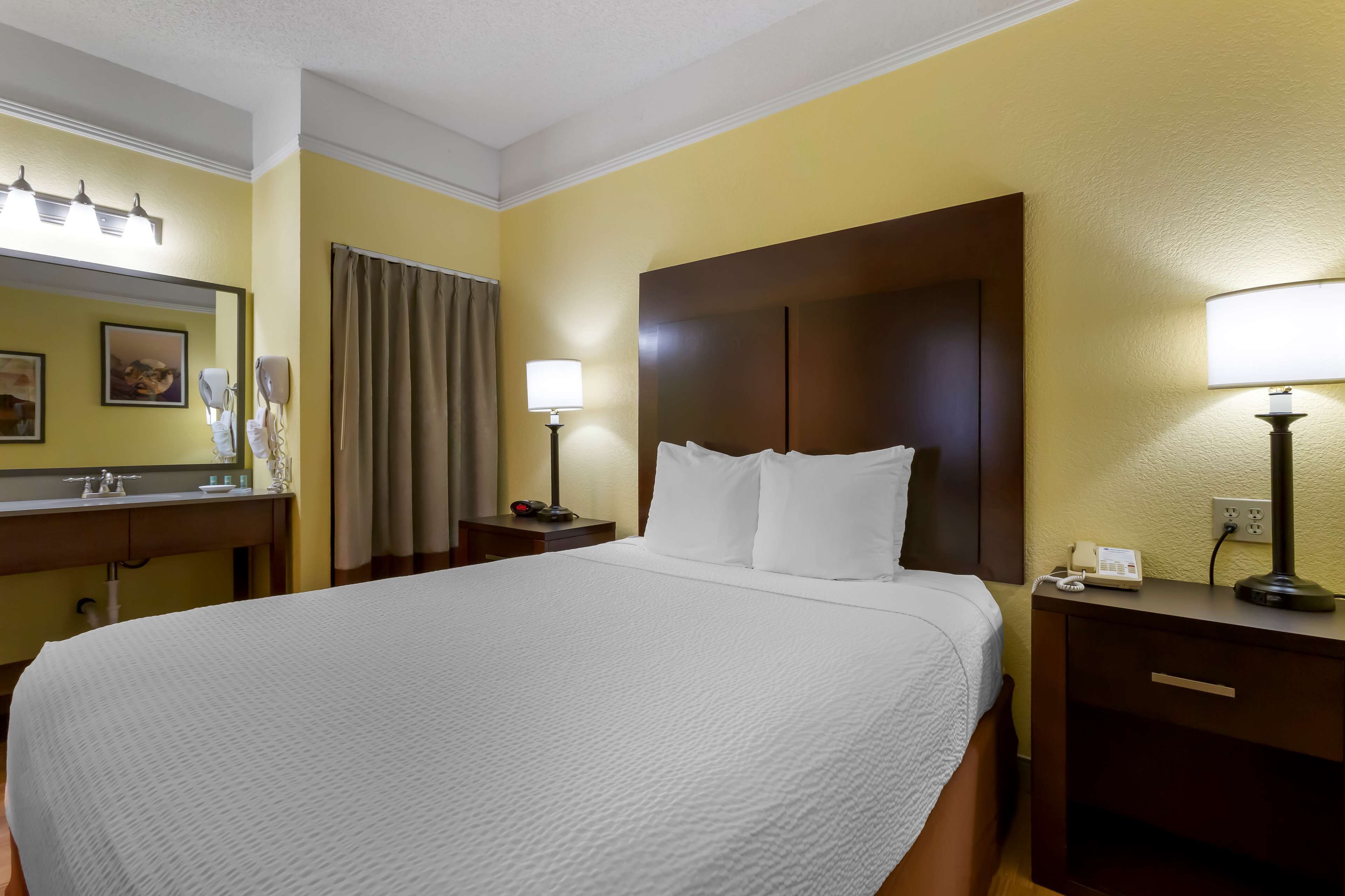 Una camera d'albergo con un letto grande, un comodino con una lampada, un tavolino con il telefono e uno specchio illuminato.