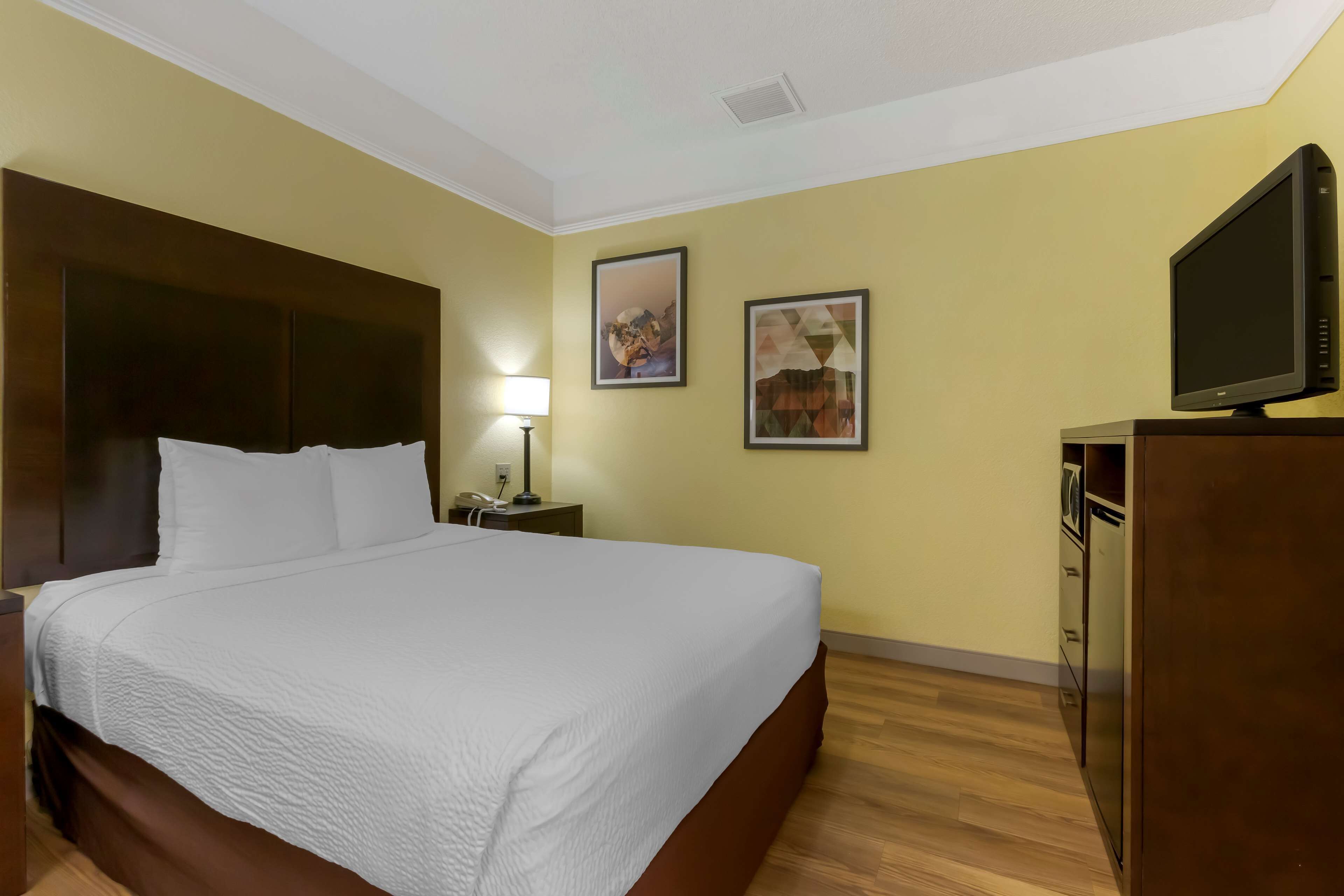 A hotel room with a large bed, a bedside table with a lamp, a TV on a dresser, and two framed pictures on the wall.