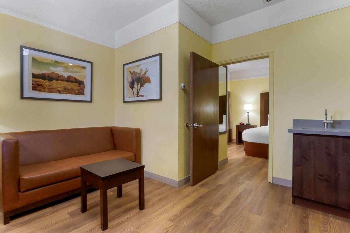 A hotel room with a brown leather sofa, a small wooden table, a bed, a desk with a sink, and two framed pictures on the wall.