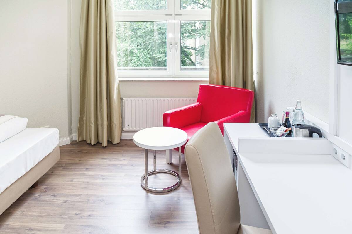 Une pièce moderne avec un canapé blanc, un fauteuil rouge, une petite table ronde et un bureau blanc sur lequel se trouve un plateau contenant des bouteilles et des verres.