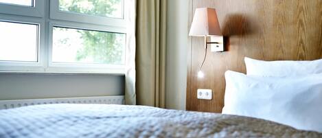 A hotel room with a bed, a window, a wall-mounted lamp, and a wooden headboard.