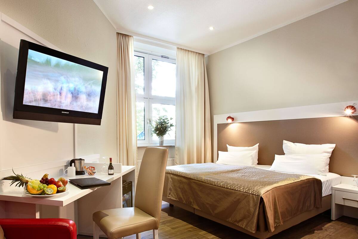 Une chambre d’hôtel avec un lit, un bureau équipé d’un ordinateur, une télévision et une fenêtre avec des rideaux.