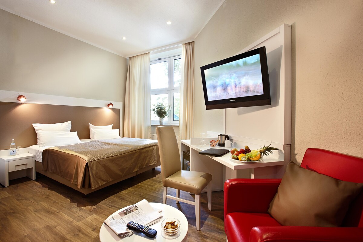 A hotel room with a bed, desk, TV, and a red chair.