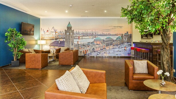 A lobby with seating areas, a large mural of a cityscape, and a tree. - Hotel Domicil Hamburg by Golden Tulip (Hamburg)