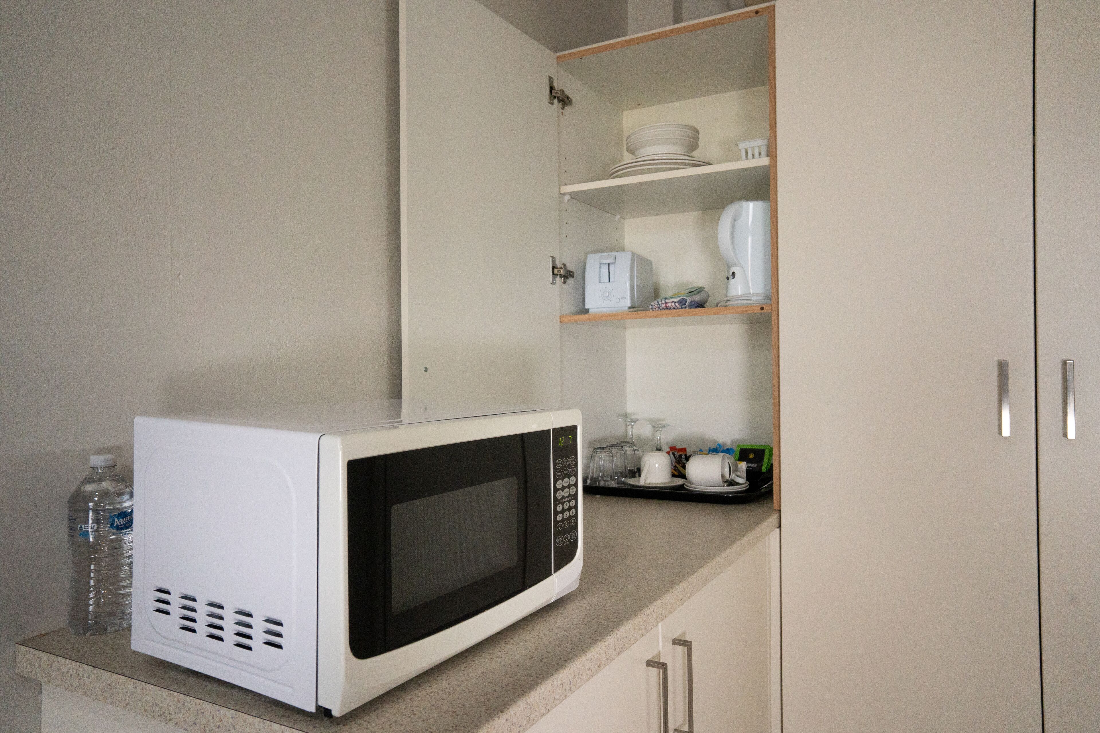 A kitchen with a microwave on the counter, a water bottle, and a cabinet with shelves containing dishes and a toaster.